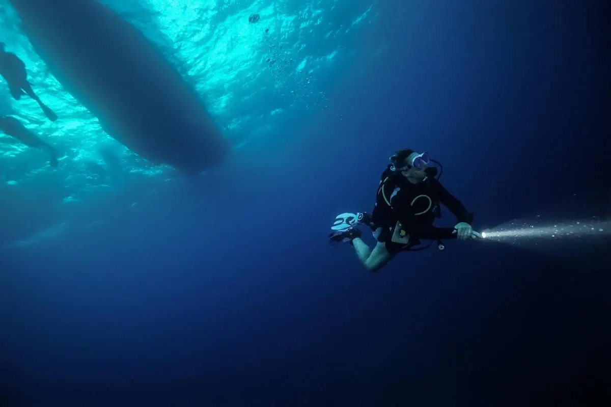 Buceador descendiendo con el haz de su linterna bajo el barco mientras se apaga la luz
