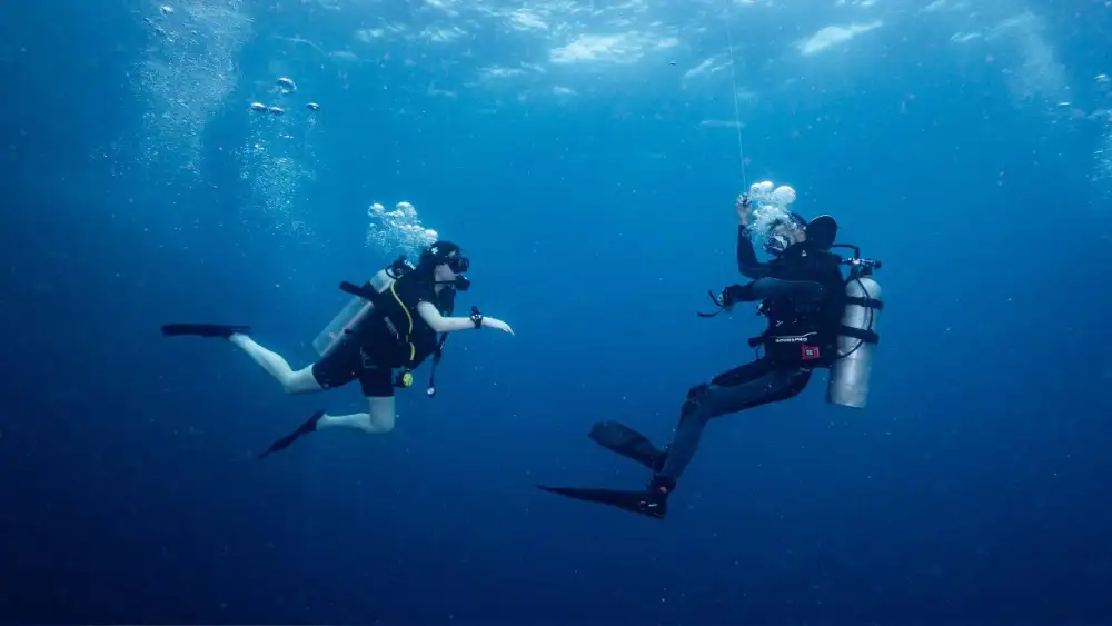 Two advanced divers exploring a deep rocky dive site in Koh Tao, one holding a torch, with soft corals on the boulders