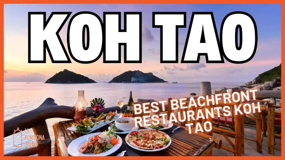 Restaurant tables set on the sand at the water's edge on Koh Tao at sunset, with longtail boats in the bay