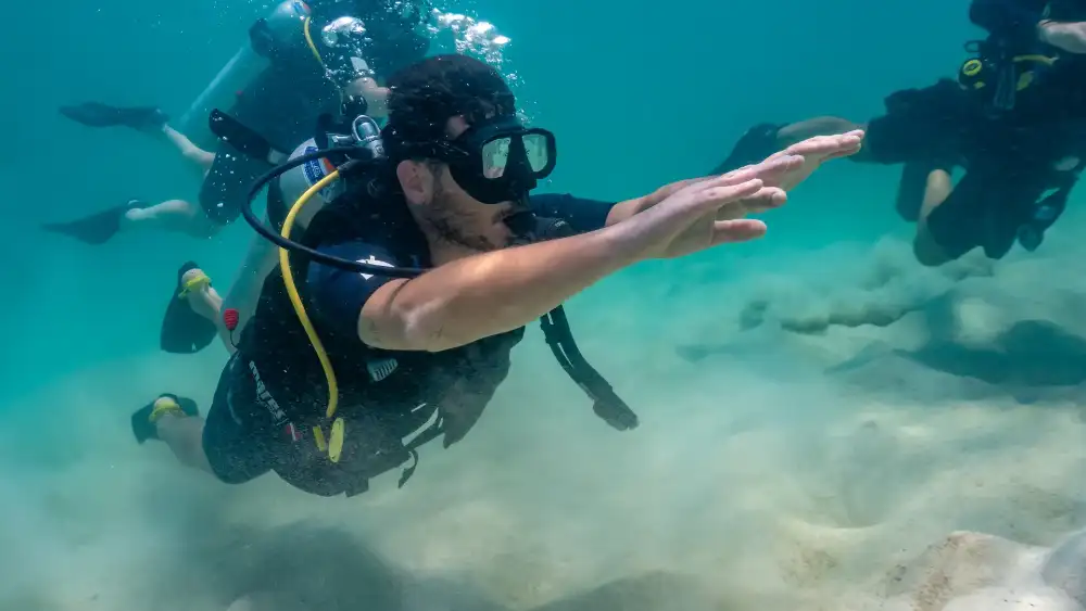 A student diver practising hovering over a sandy bottom in shallow water, with other divers behind