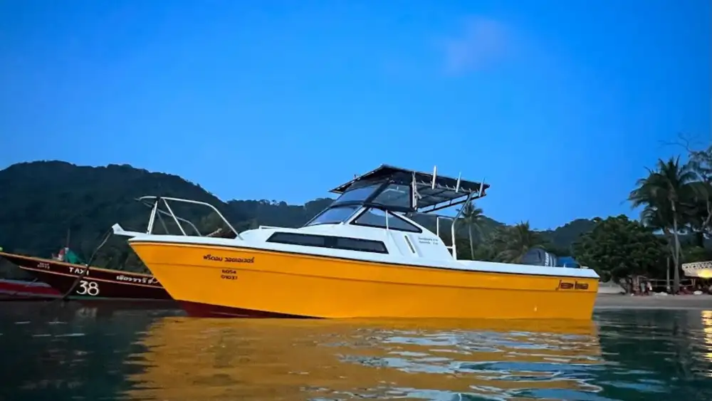 A yellow and white sport-fishing speedboat with a shade canopy moored in calm water at dusk, with a longtail taxi boat and jungle-covered hills behind