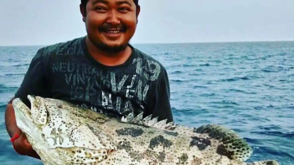 A smiling Thai fisherman on a boat holding a large spotted grouper with both arms, open sea behind him