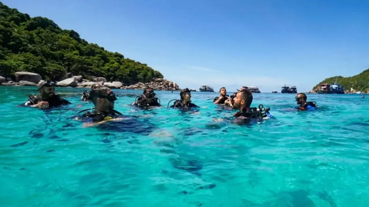 A group of divers floating at the surface in turquoise water off Koh Tao, dive boats and the island's green hills behind them
