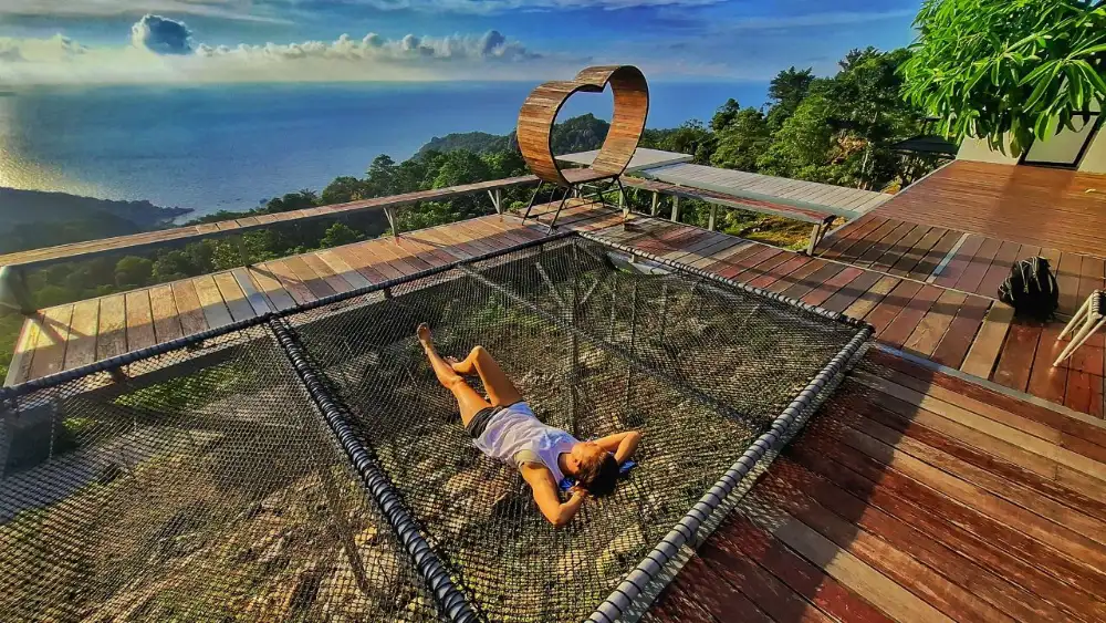 A woman lying on a net platform at a hilltop viewpoint on Koh Tao, with a heart-shaped wooden frame, wooden decking and the sea and jungle below