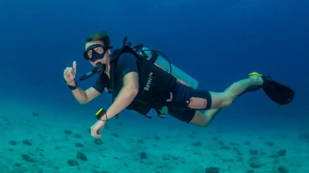 A scuba diver hovering above a sandy bottom in clear water at Koh Tao, signalling to the camera