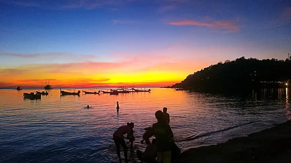 Sunset over a bay on Koh Tao with longtail boats at anchor and people silhouetted on the shore