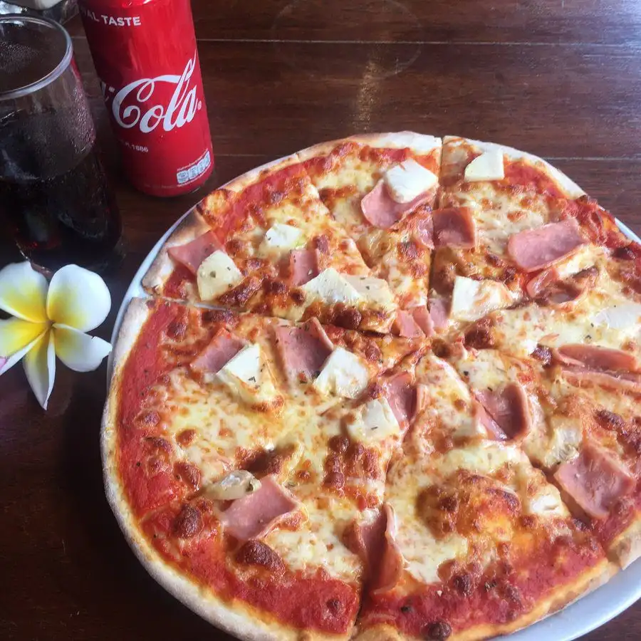 A ham and cheese thin-crust pizza on a wooden table next to a can of cola and a frangipani flower