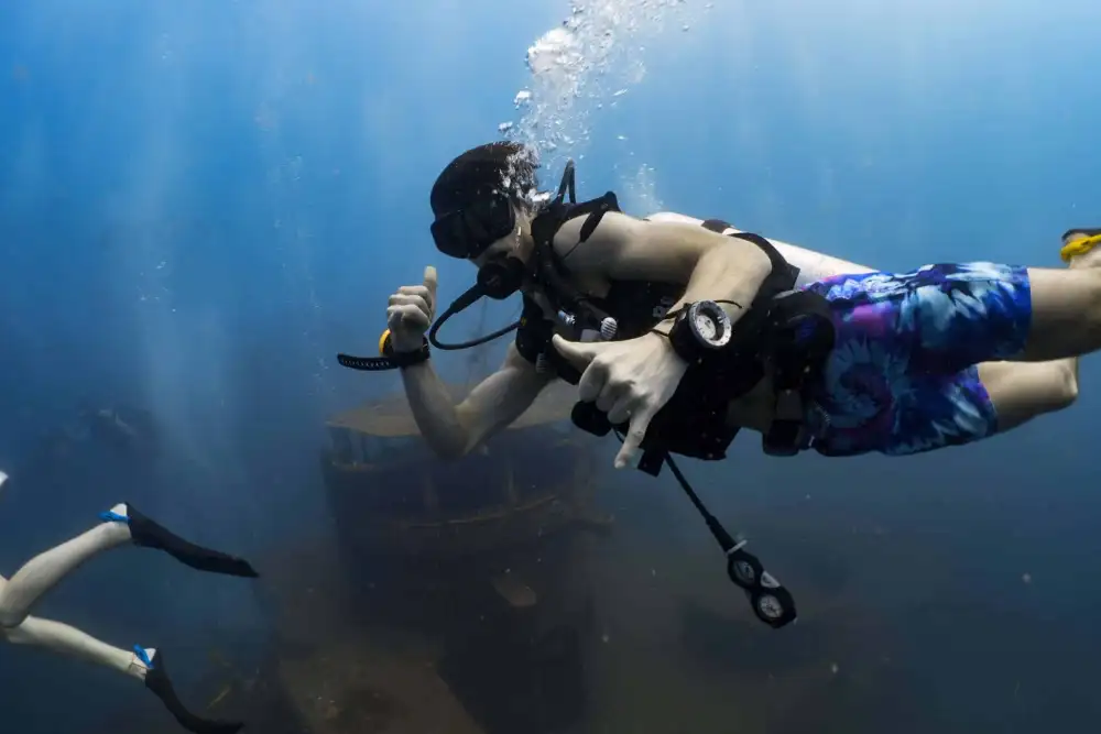 A scuba diver in board shorts giving a shaka sign above a wreck in blue water off Koh Tao, another diver's fins in the foreground