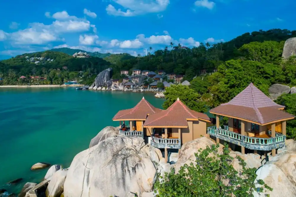 Resort bungalows with red-tiled roofs perched on granite boulders above a turquoise bay on Koh Tao, green hills behind