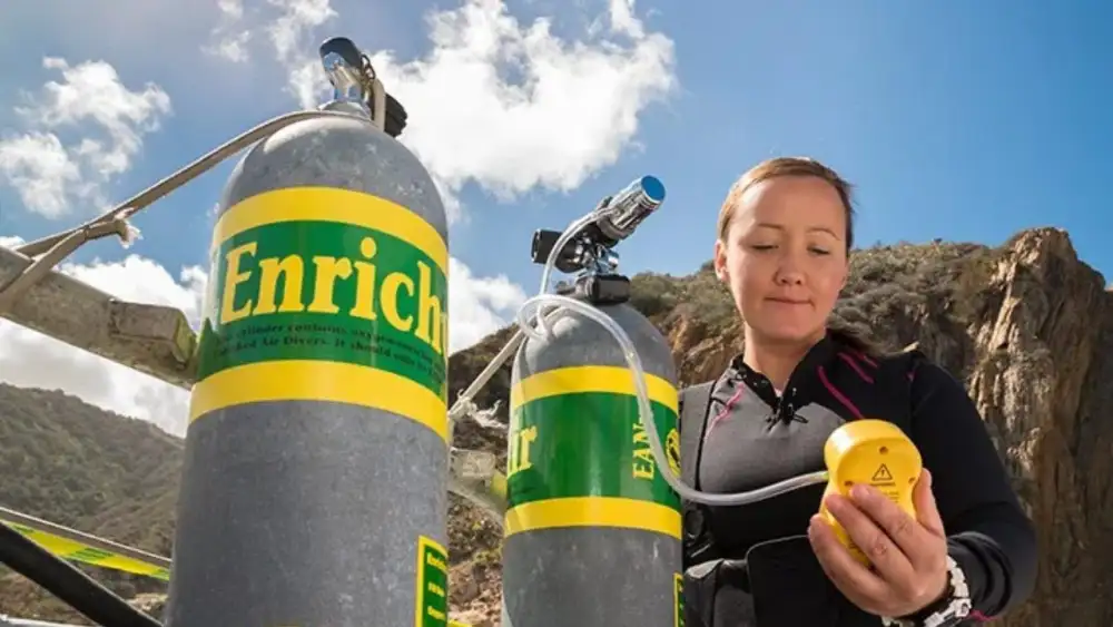 A diver holding a yellow oxygen analyser against the valve of a nitrox tank to check the gas mix before a dive, sea and rocks behind her