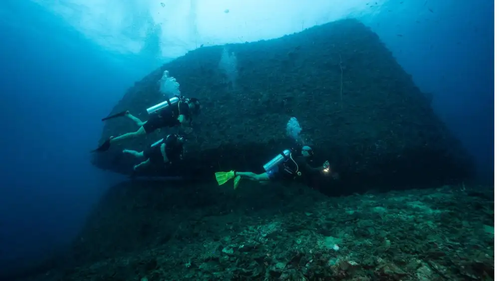 A diver kneeling on the sandy bottom practising slow, controlled breathing with the instructor watching