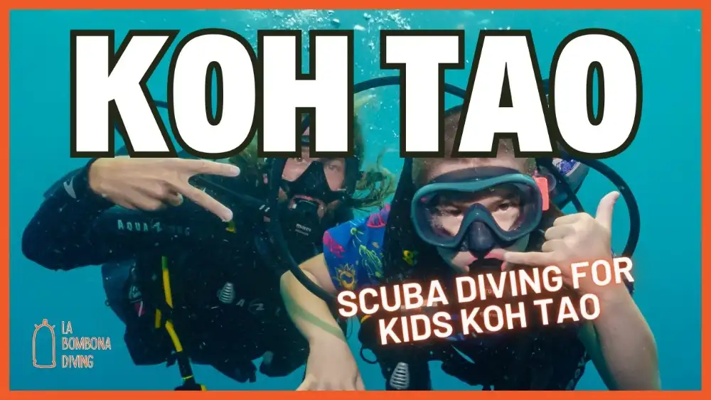 A young diver in a mask and wetsuit smiling at the surface next to an instructor in the calm, shallow water of a Koh Tao bay