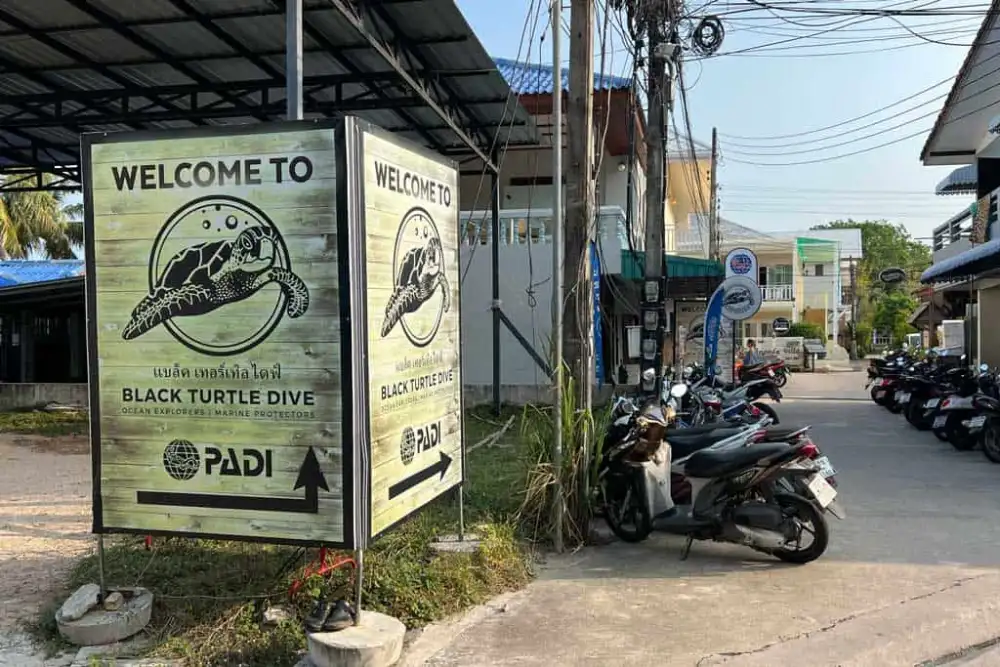 The wooden 'Welcome to Black Turtle Dive' sign with a turtle logo and PADI arrow at the roadside, with scooters parked along the street on Koh Tao