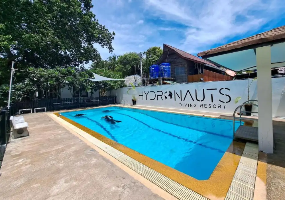 The training pool at Hydronauts Diving Resort on Koh Tao, with two divers practising underwater and the resort's name painted on the white wall behind